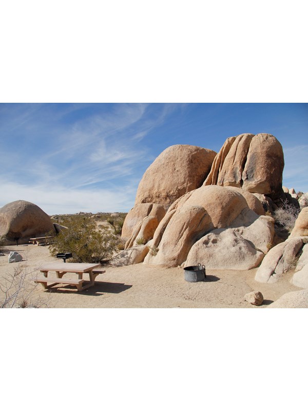 A picnic table and fire pit are in a campsite. Behind them are large boulders.