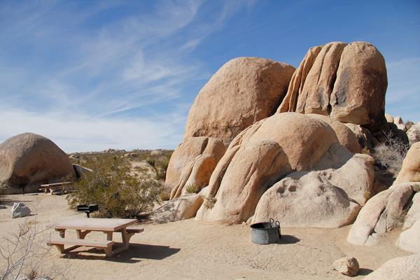 A picnic table and fire pit are in a campsite. Behind them are large boulders.