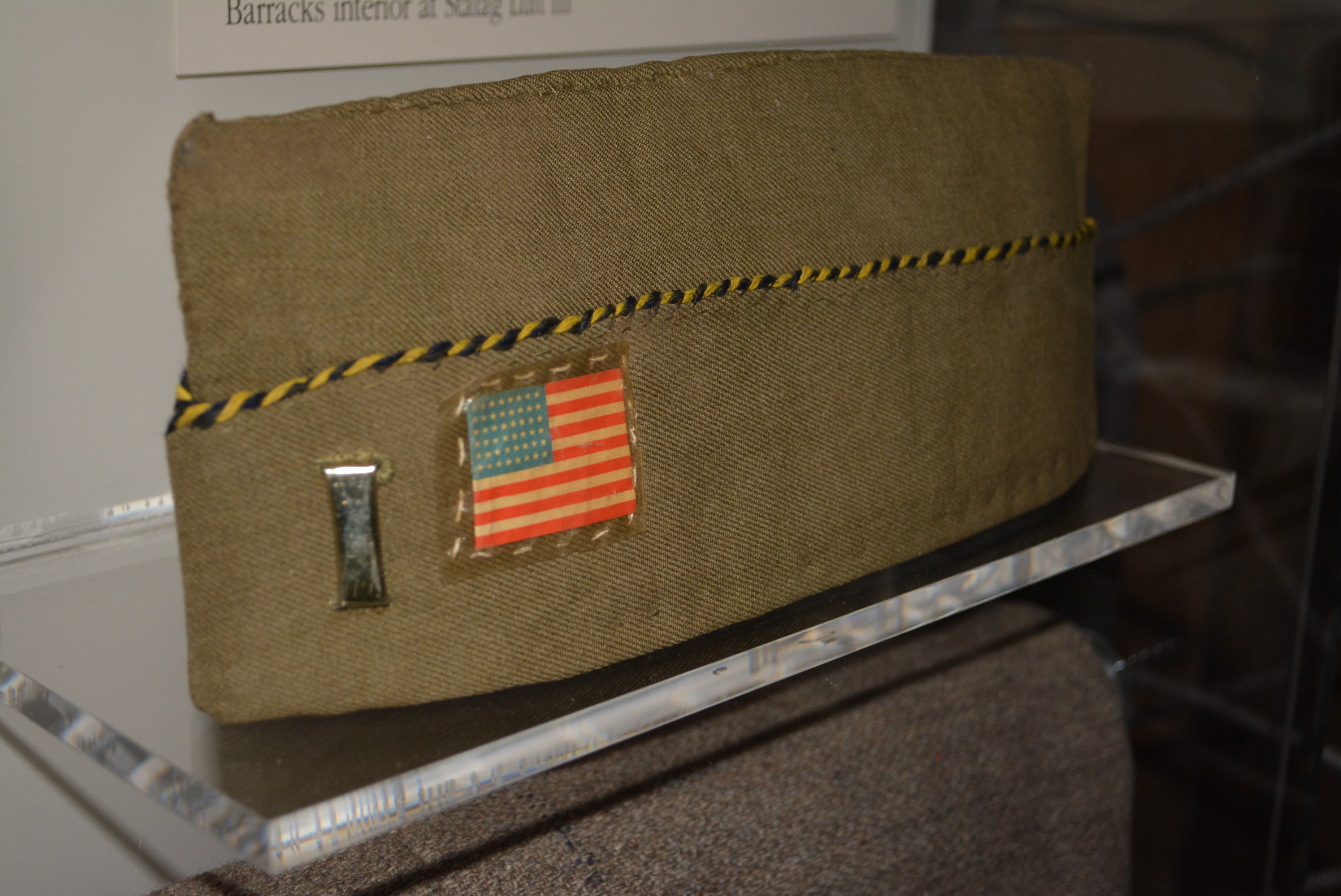 A military garrison cap with a small U.S. flag and silver bar in an exhibit case