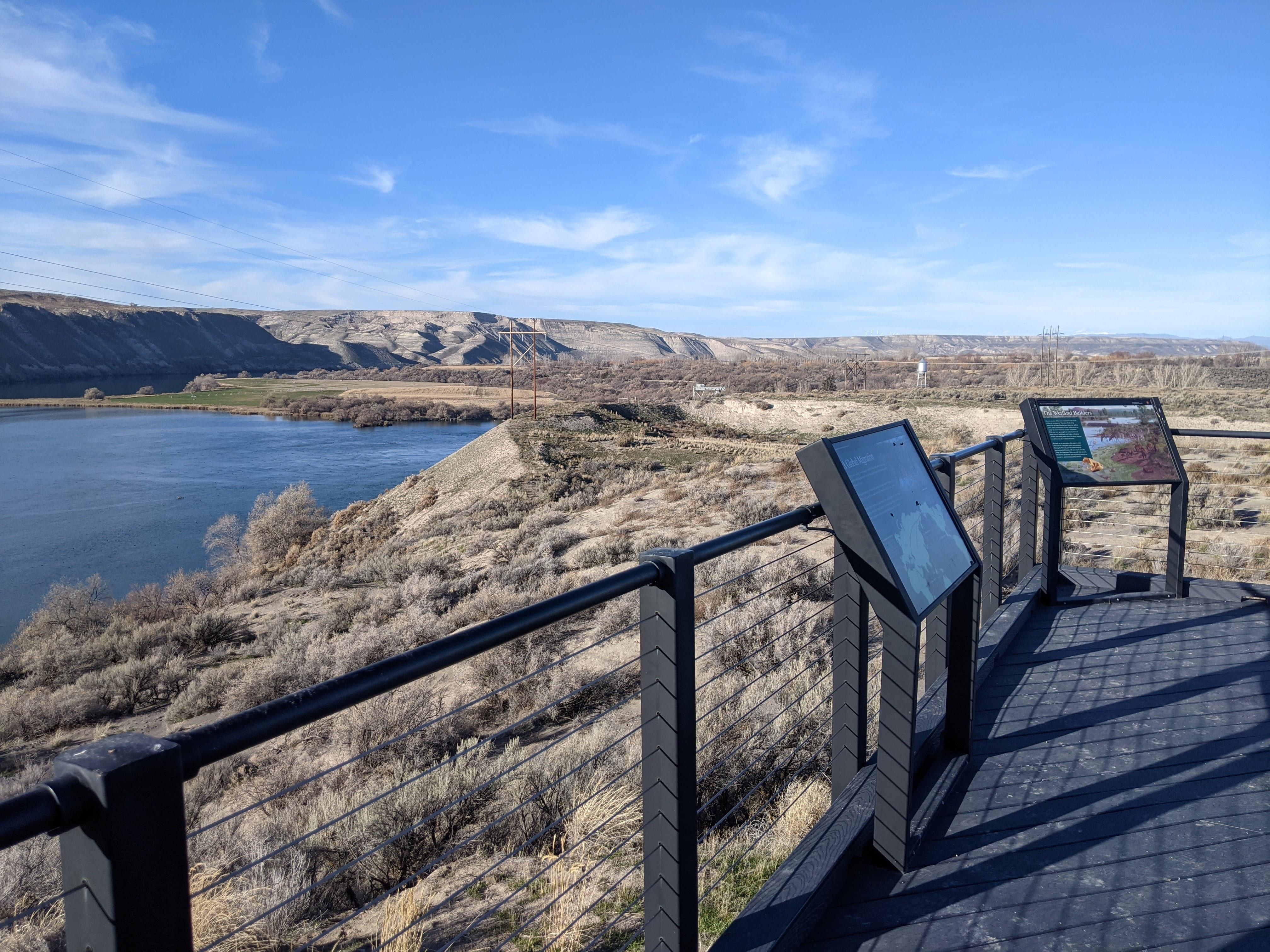 A wooden platform with railing, along with two exhibit signs, overlooks the river and fossil beds.
