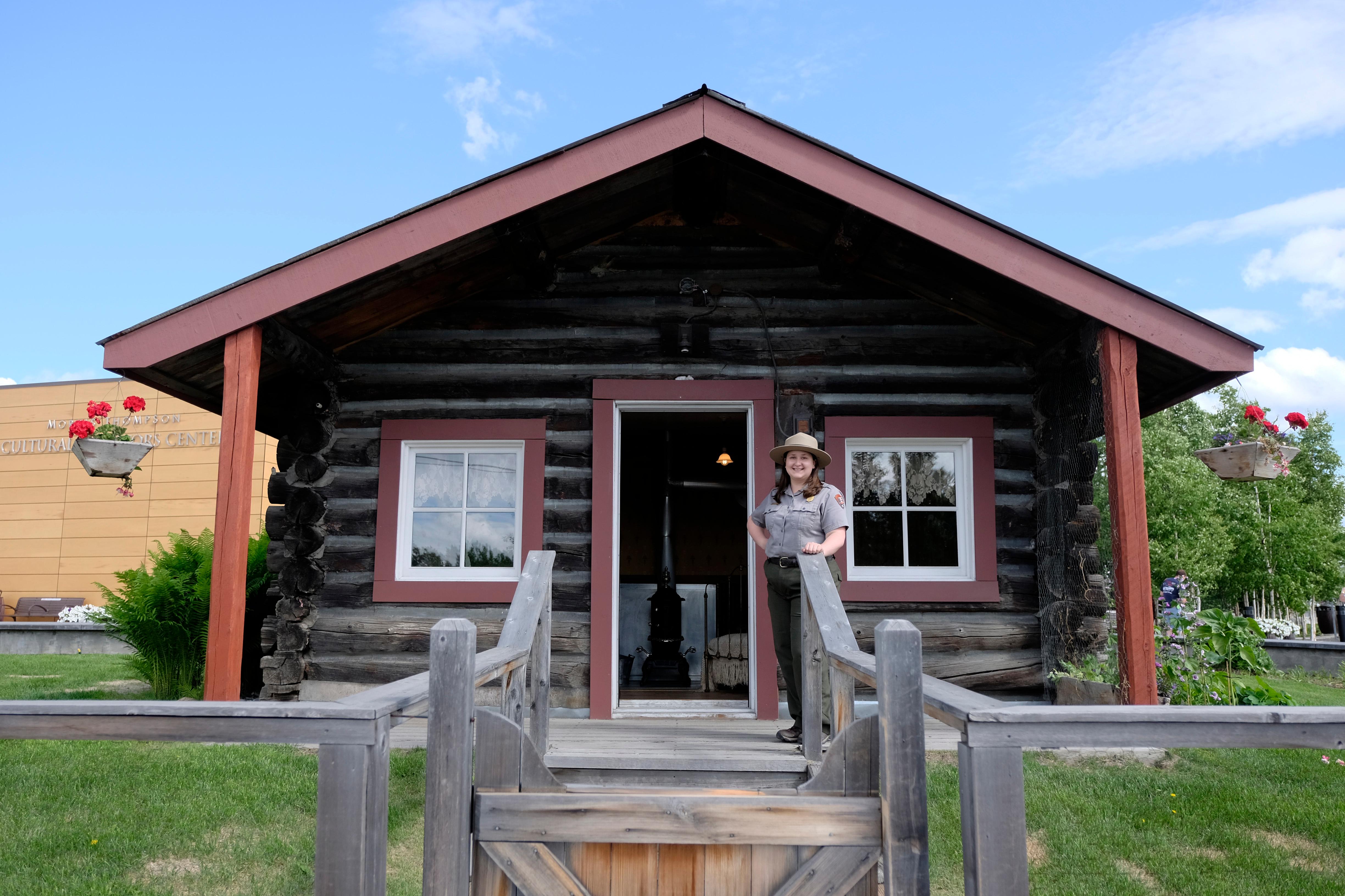 A national park ranger stands outside the open door of a historic log cabin