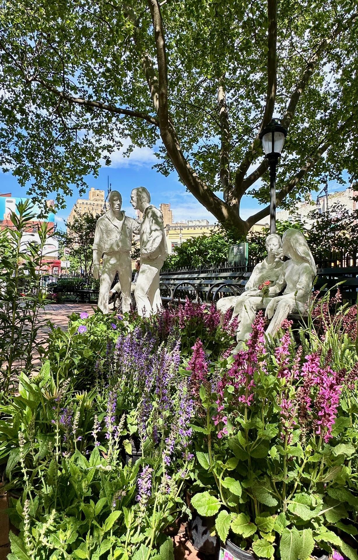 Pink and purple flowers, behind them are four white statues, 2 men standing and 2 women sitting down