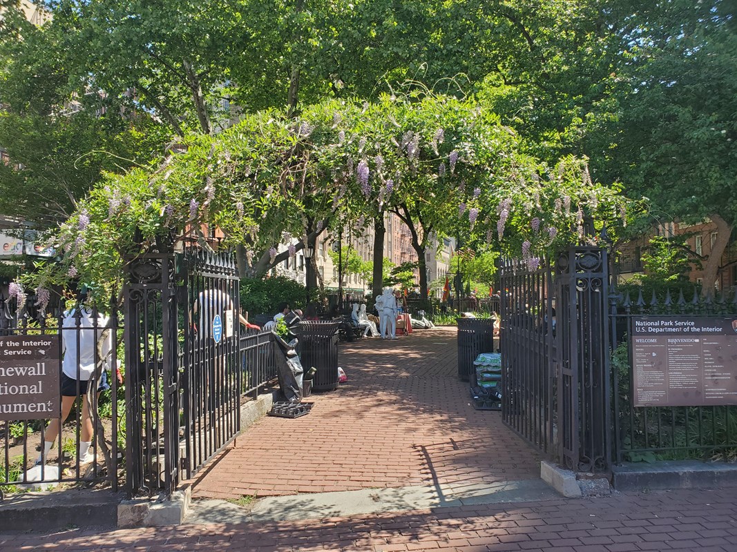 An open black fence gate with green plants blooming above it.