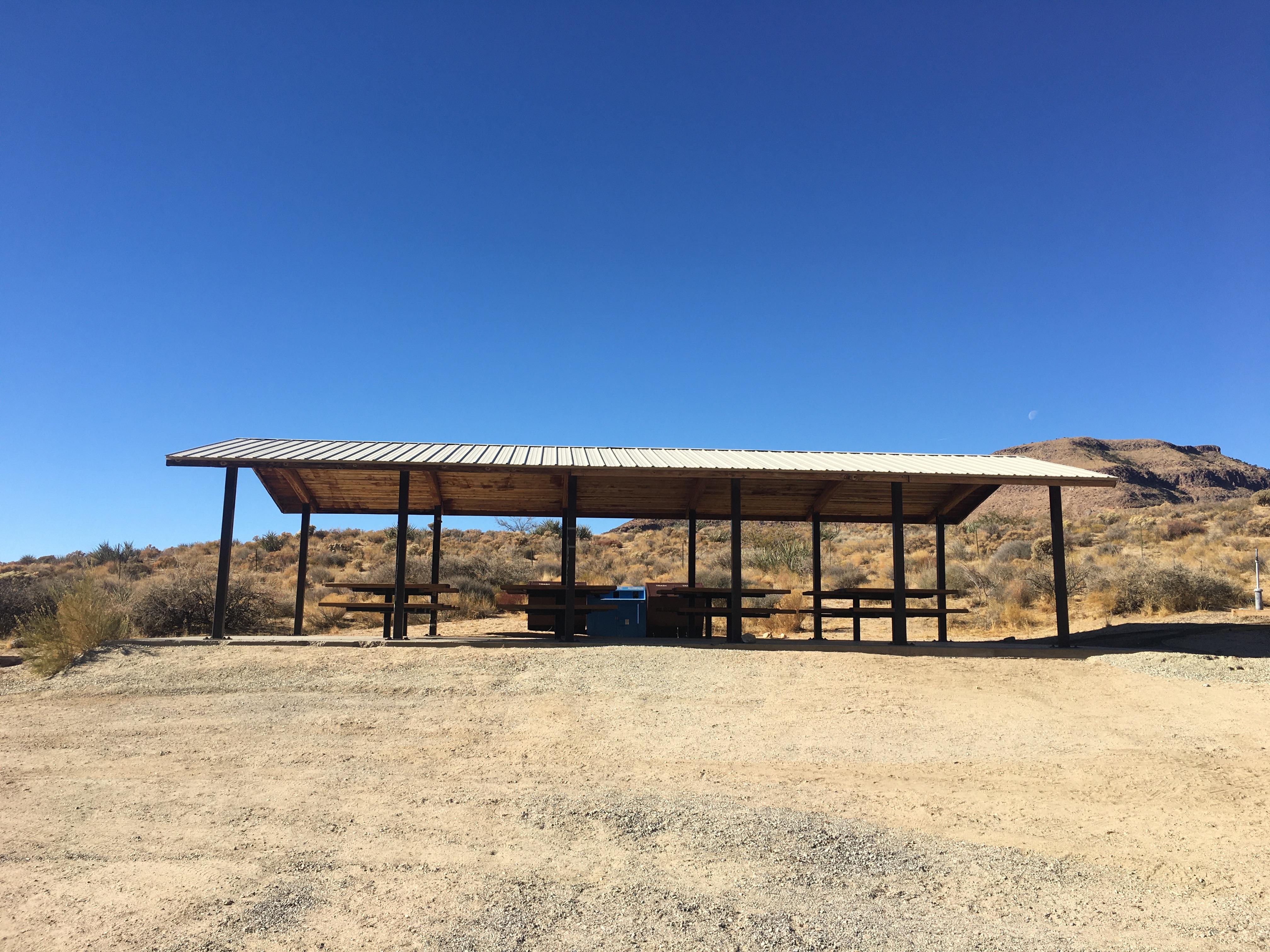 A shade structure consisting of beams holding up a solid roof. Underneath are six picnic tables.