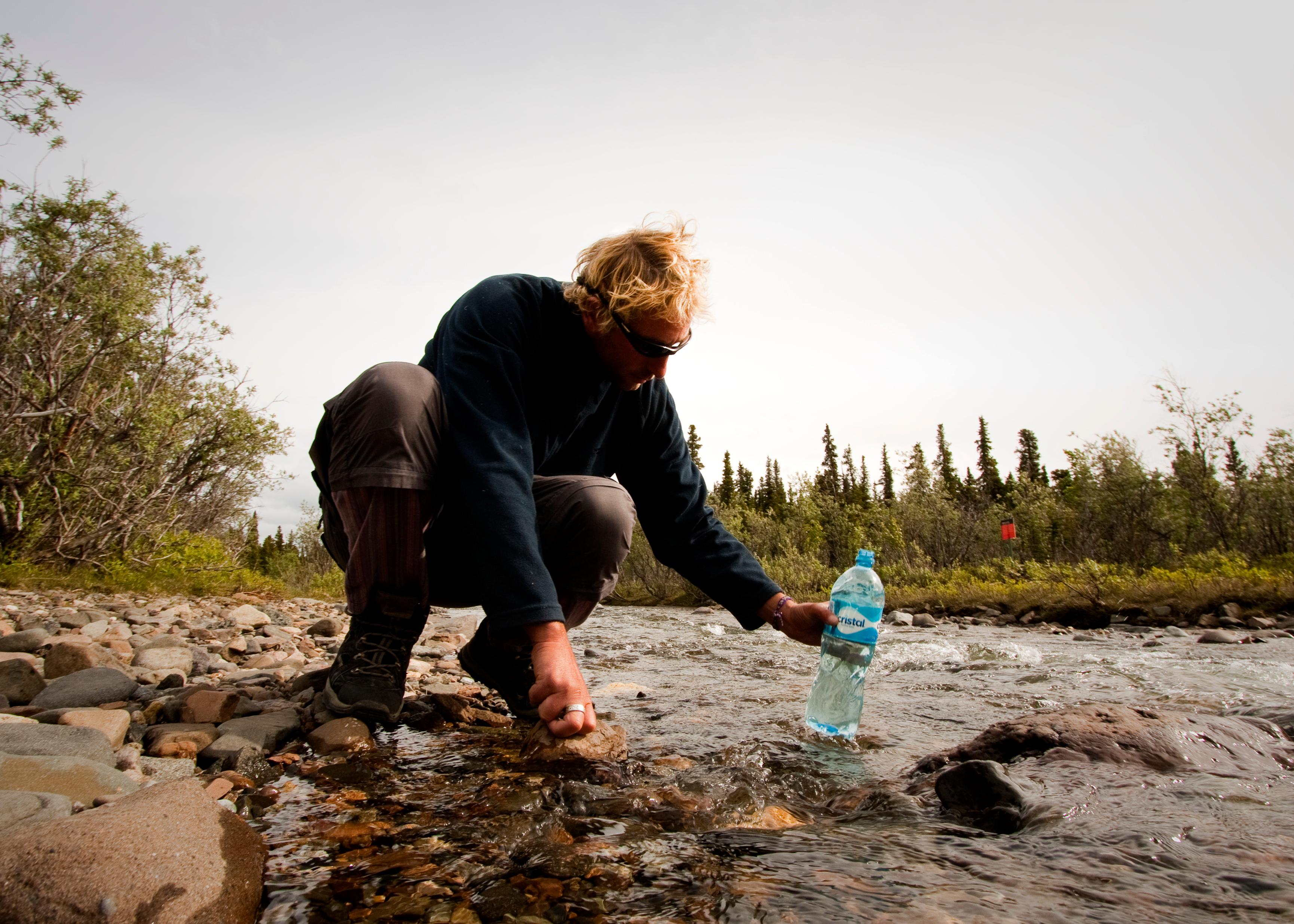 a woman filtering water into a plastic bottle from a shallow creek