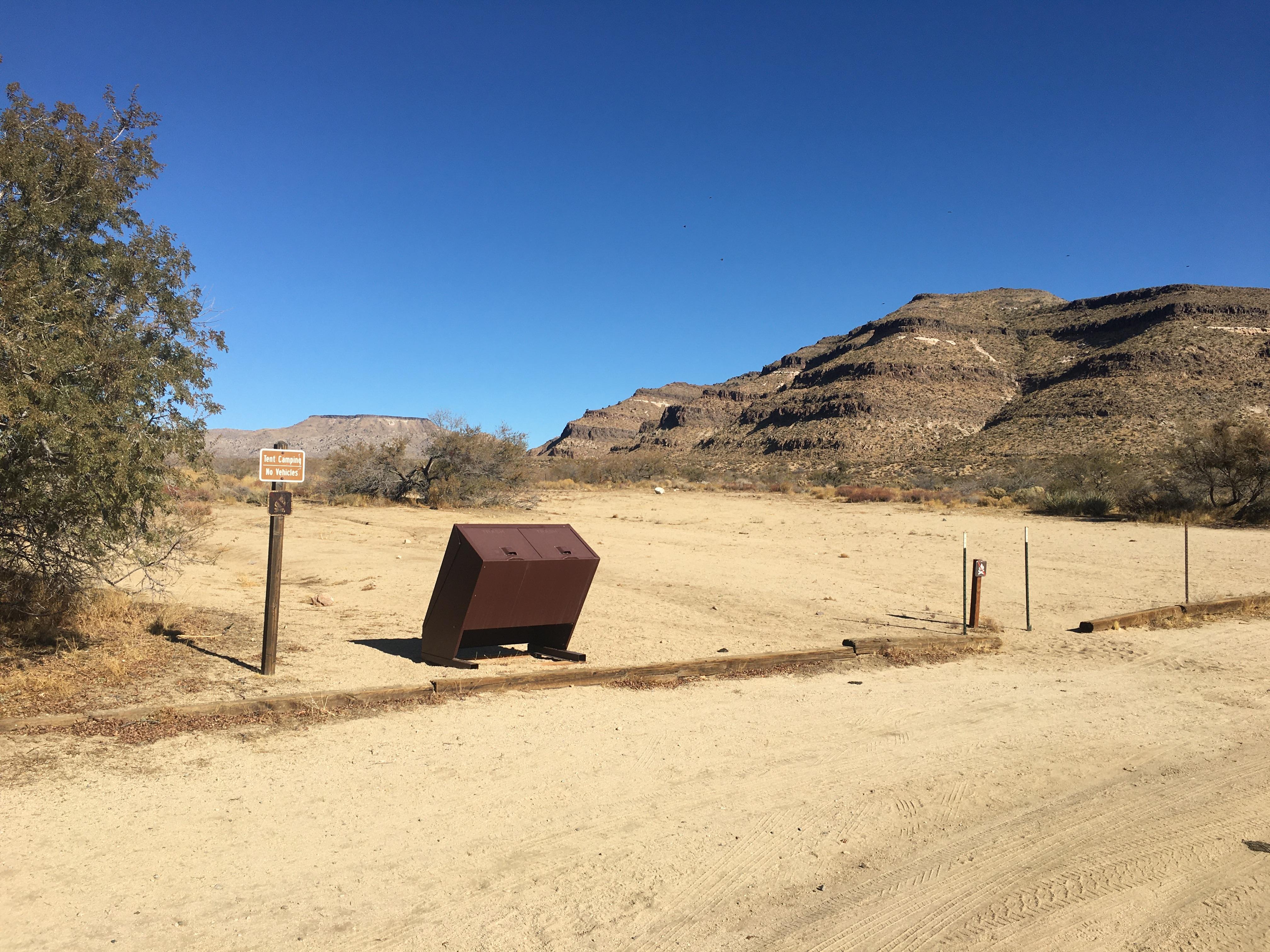 A garbage receptacle at a small sign that says "tent camping, no vehicles" stand in a cleared area