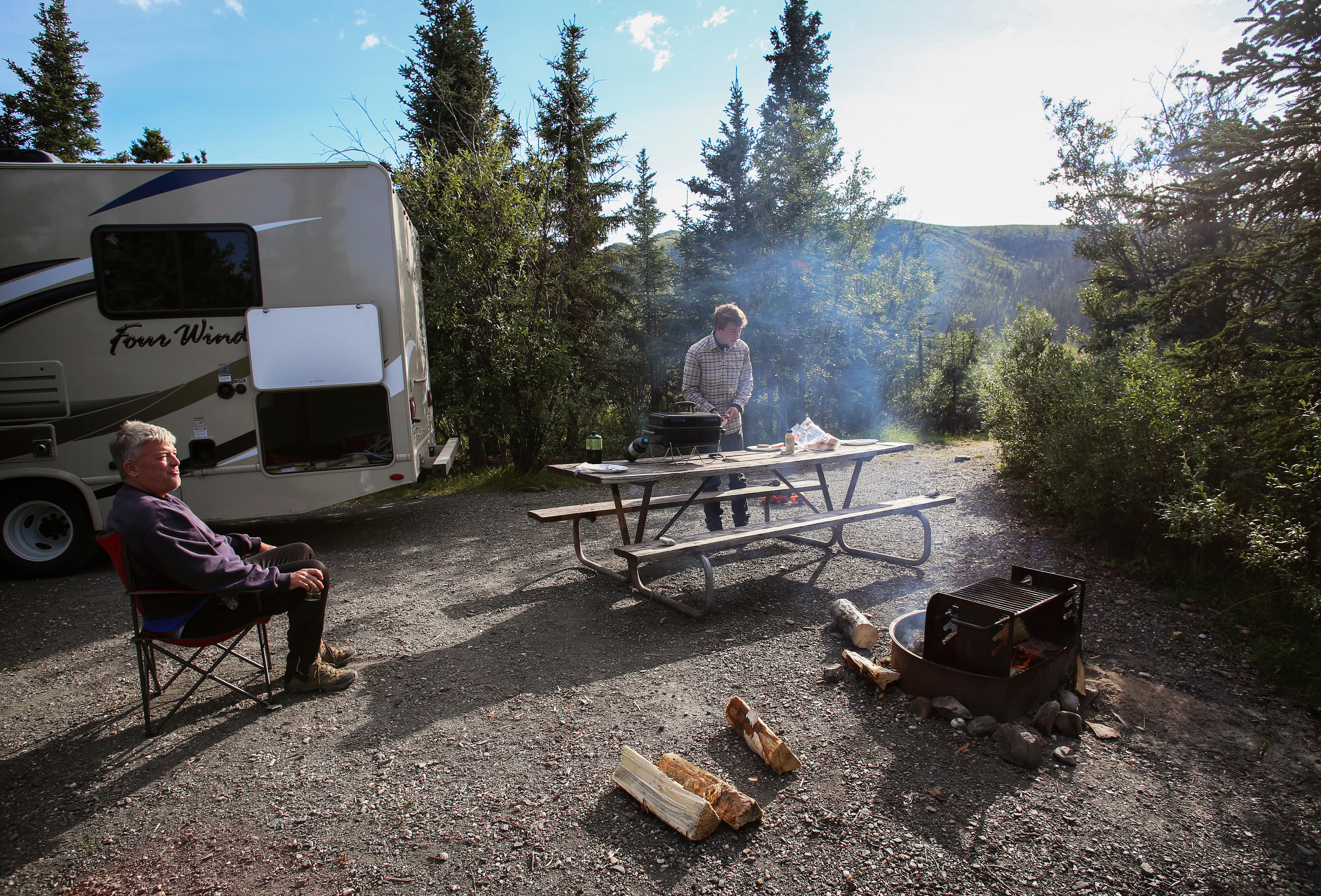 two people outside of an RV, one relaxing in a camp chair by a fire while another prepares a meal