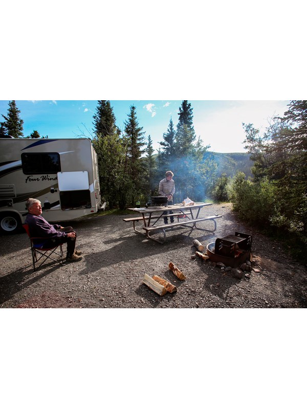 two people outside of an RV, one relaxing in a camp chair by a fire while another prepares a meal