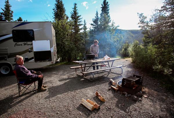 two people outside of an RV, one relaxing in a camp chair by a fire while another prepares a meal
