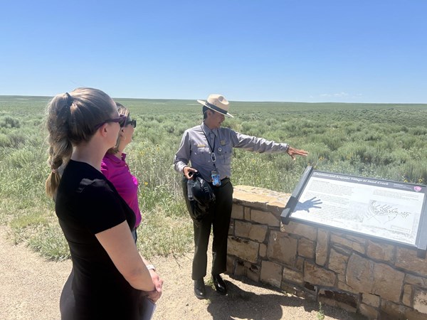 A park ranger points in the distance as two visitors stand before an outdoor exhibit panel.