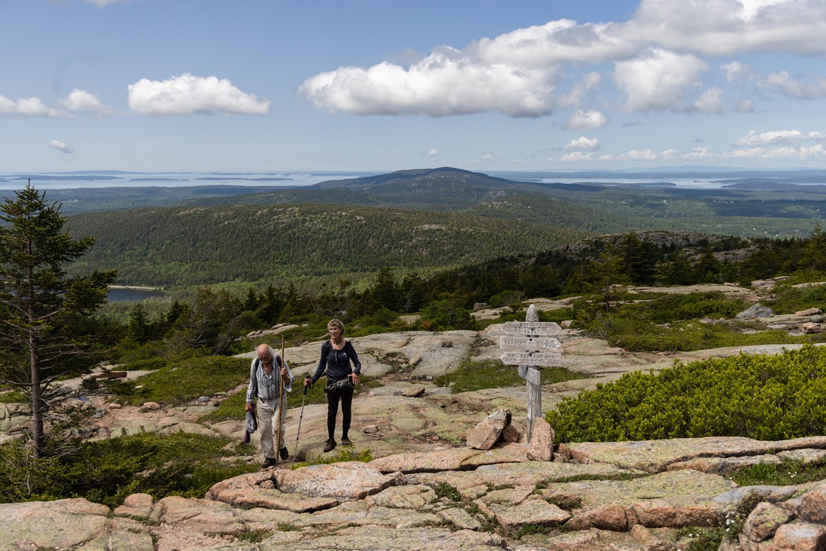 A man and woman holding walking sticks hike up a mountain summit near a wooden trail sign.
