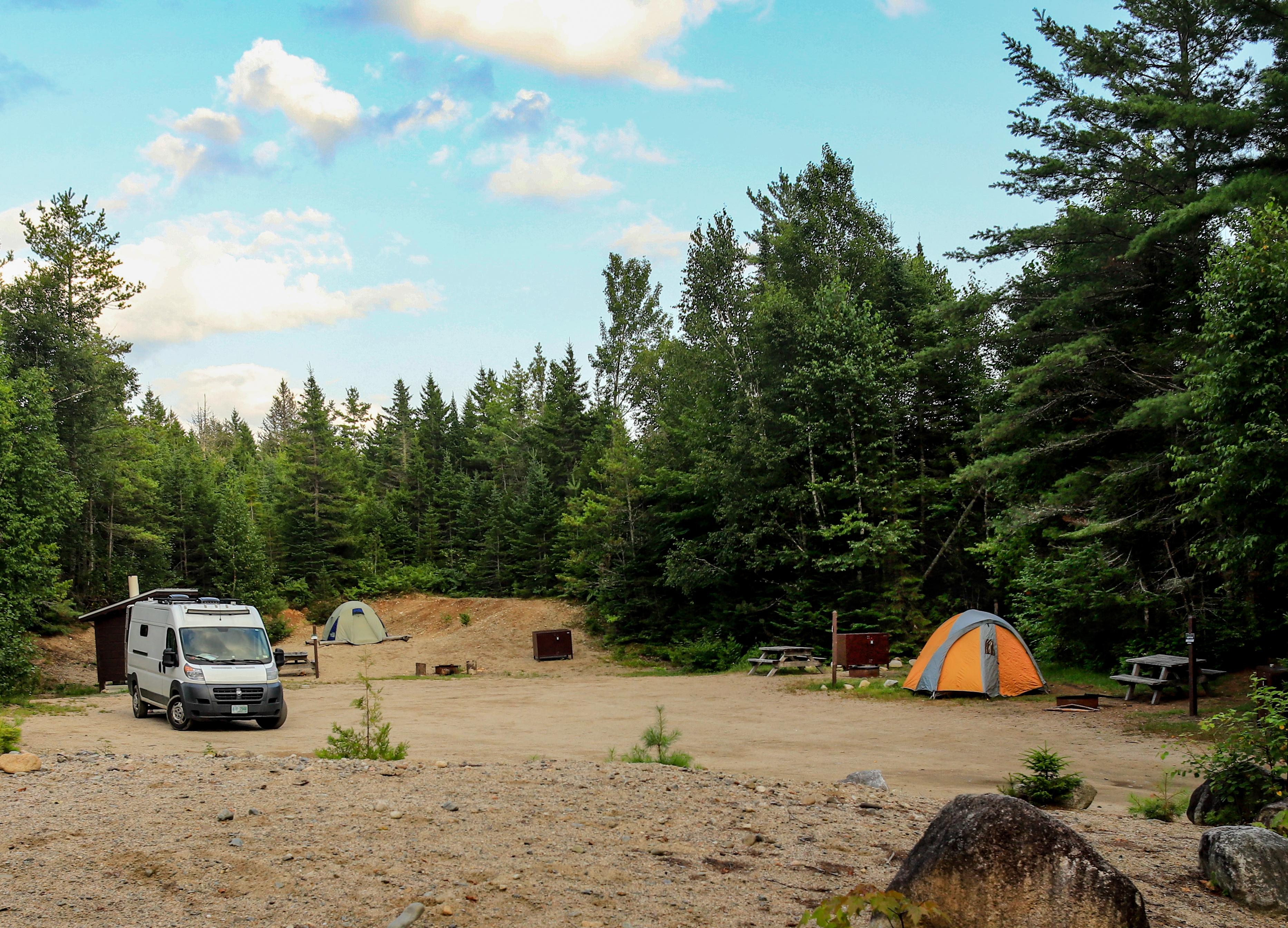 Two tents and a van set up in a flat sandy area surrounded by trees