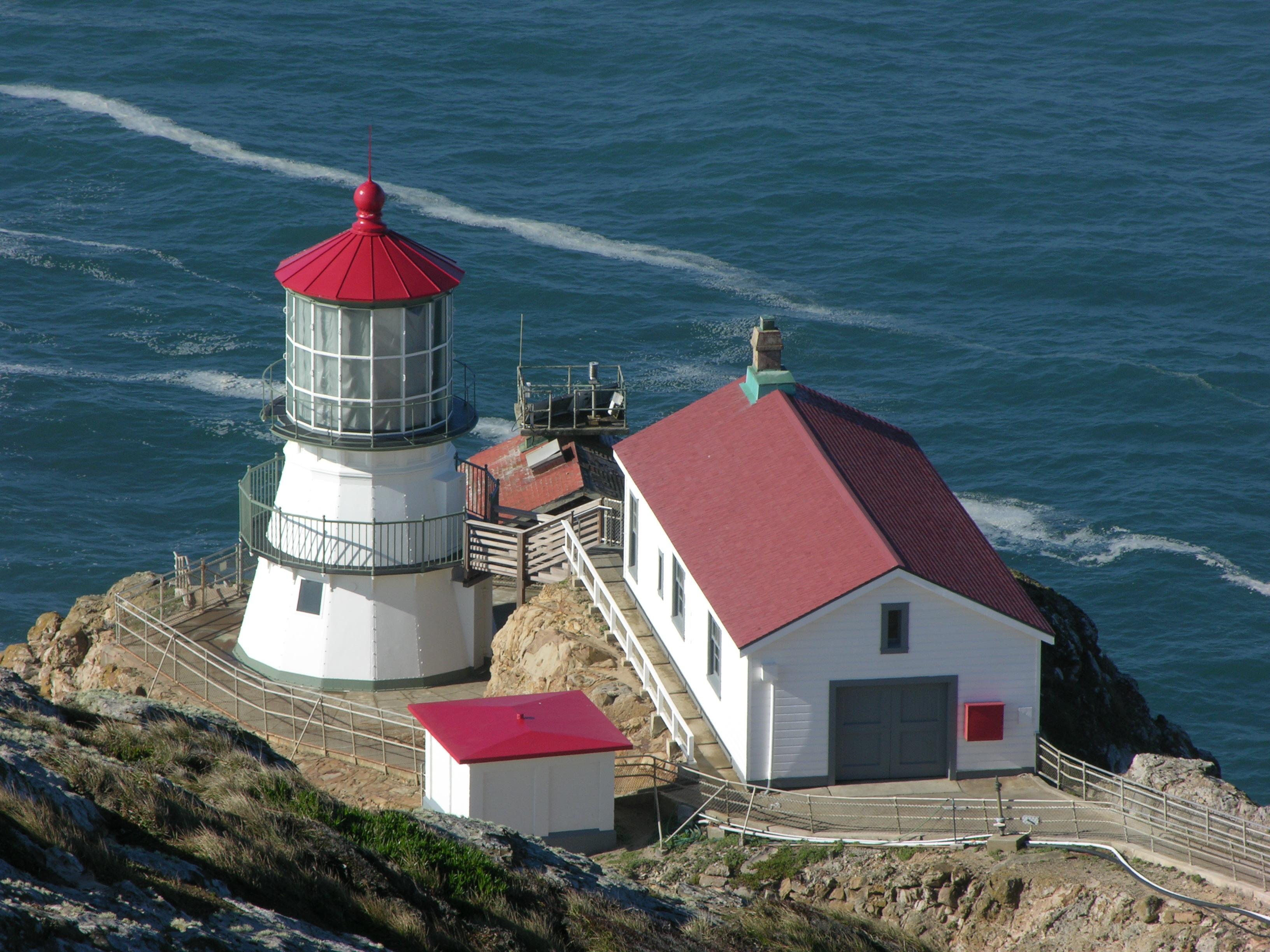 A three-story-tall, white-sided, red-roofed lighthouse adjacent to three other small buildings.