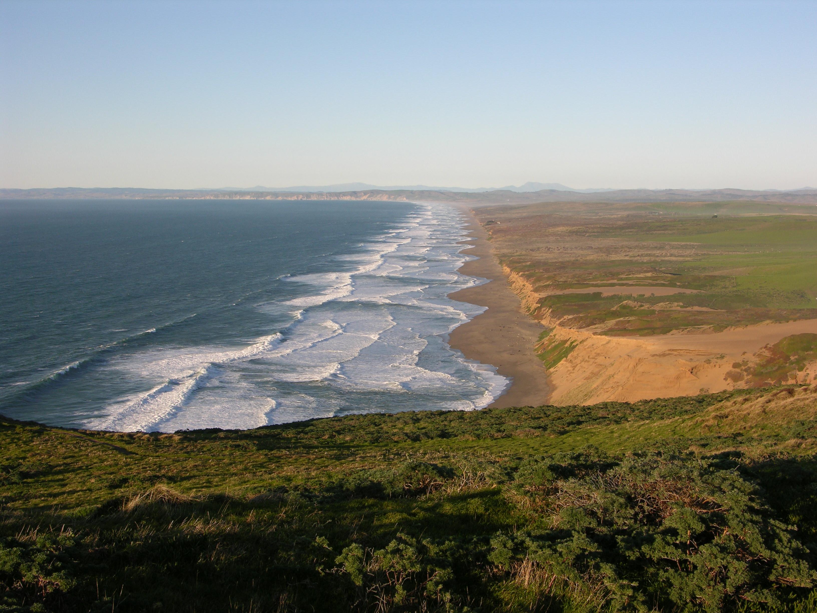 Waves wash in from the left onto a long, straight stretch of undeveloped, low-lying coastline.