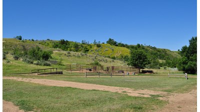 A horse corral area with green hills in the background and a gravel drive in the foreground.