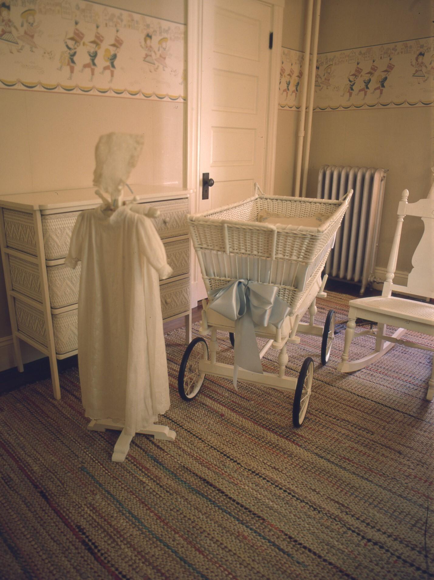 Room with white wicker furniture, including bassinet and rocking chair. Long white baby dress hangs.