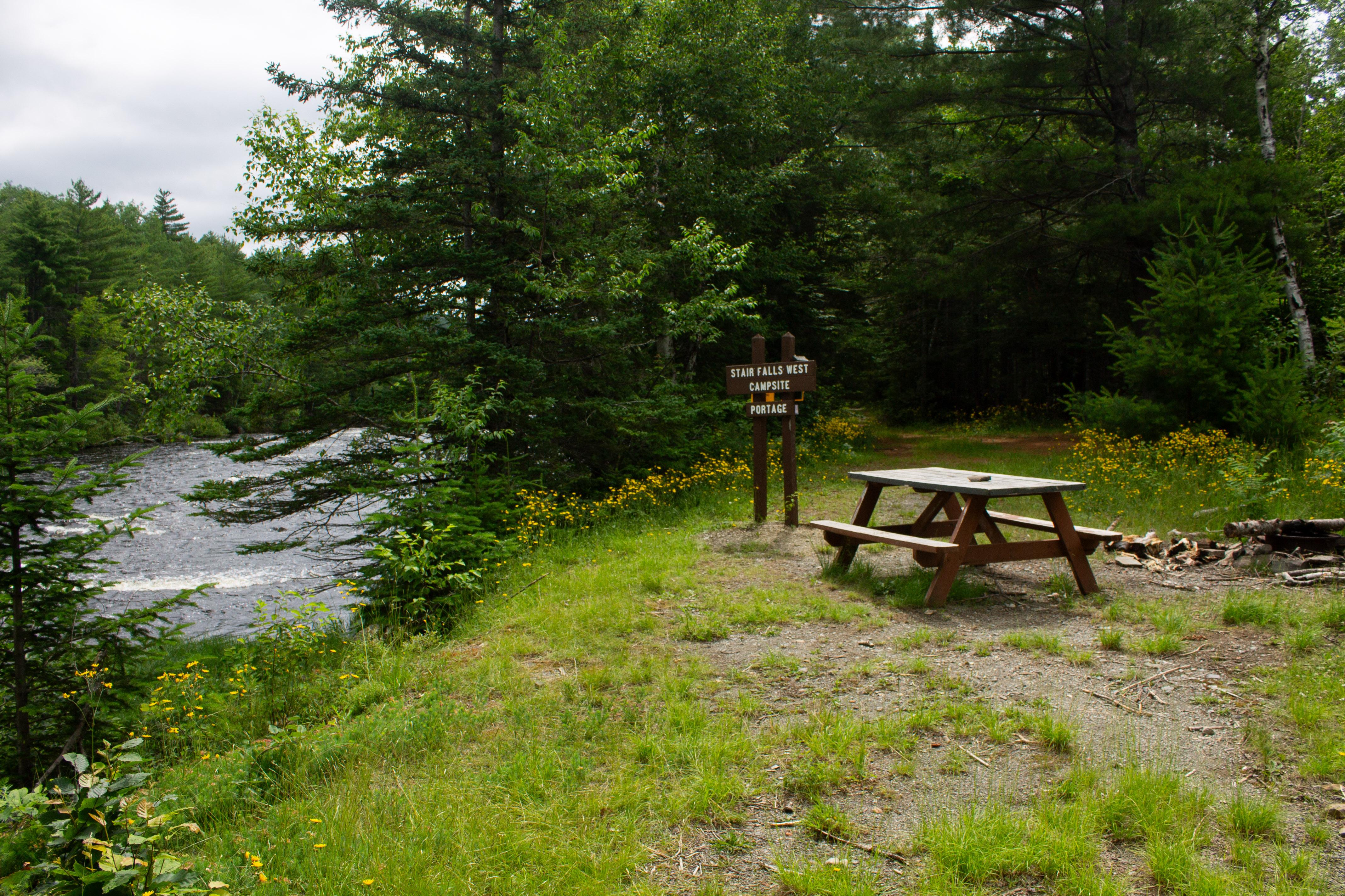 Sign reading "Stair Falls West Campsite Portage" next to a river, with a picnic table.