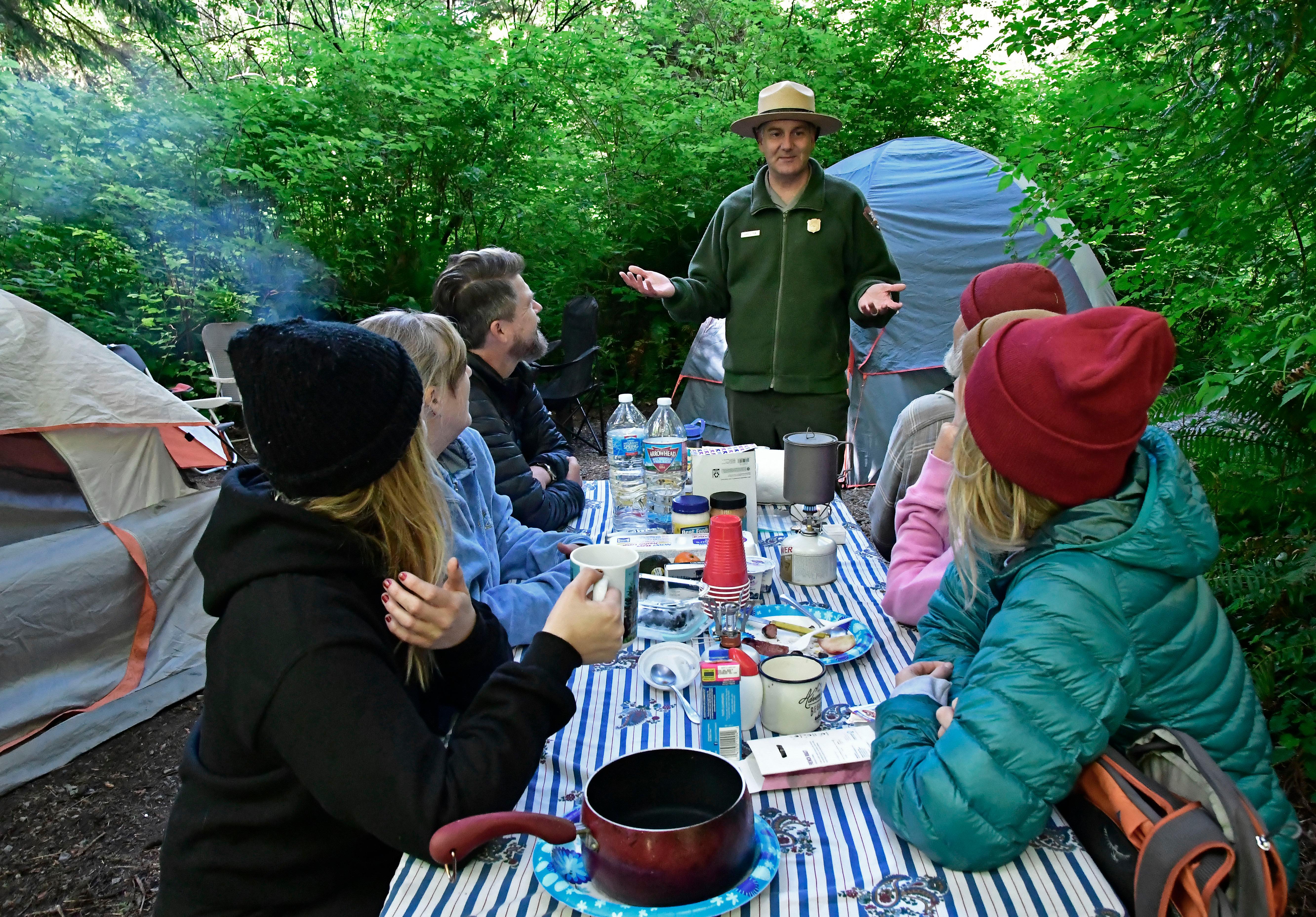 A park ranger chats with a  family sitting at a picnic table. Two tents are nearby.