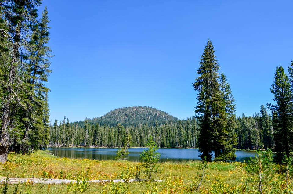 A blue lake fronted by green and yellow meadow, lined by conifers, and backed by a round, volcanic p