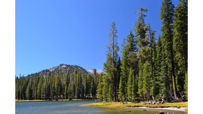 People sit and stand on a grassy shore of a mountain lake.