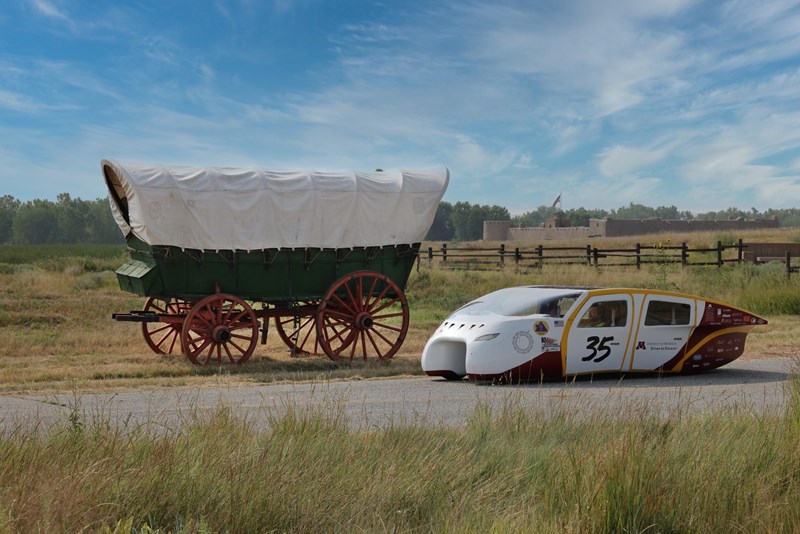 The Past and Future at Bent's Old Fort A Conestoga wagon and a solar race car stand on a road with a large adobe fort in the background