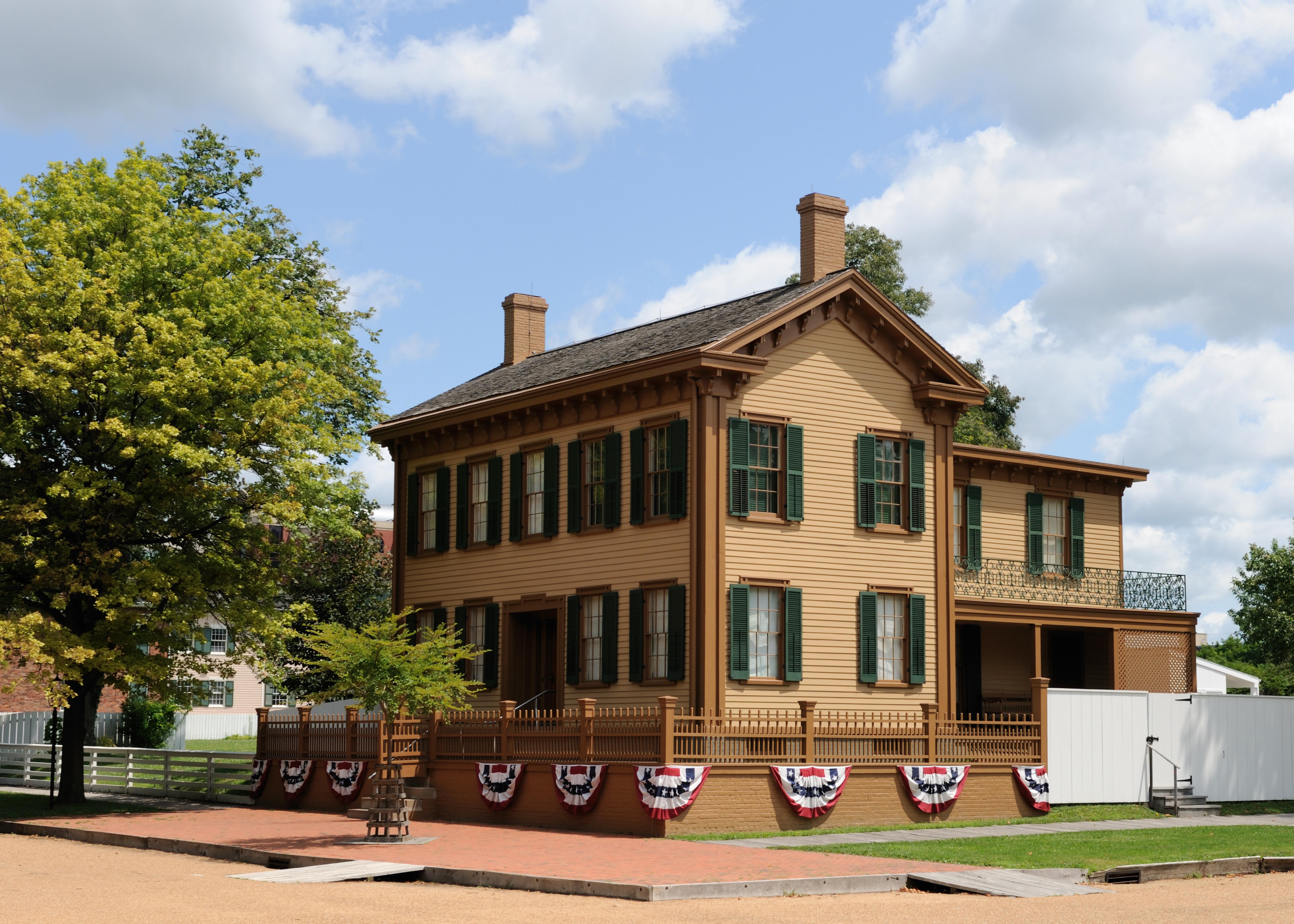 2 story tan house with green shutters. Red, white, and blue striped bunting draped below its fence