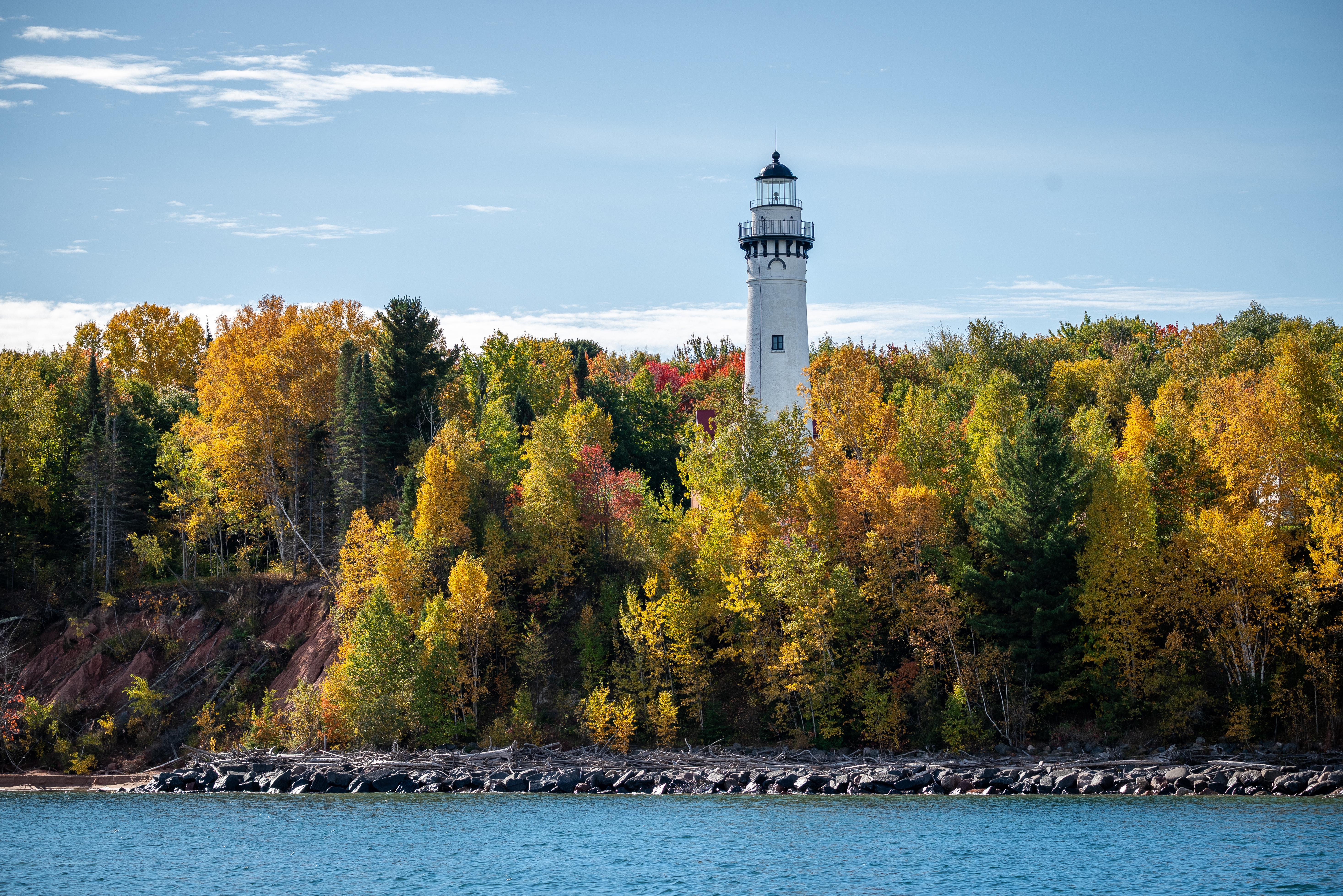 White lighthouse rising above colorful fall trees along a rocky shoreline under a blue sky.