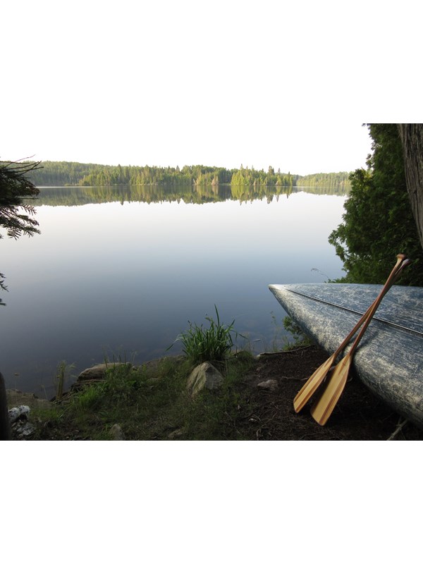 Calm waters of a small lake greet the bow of an overturned canoe with paddles resting against it.