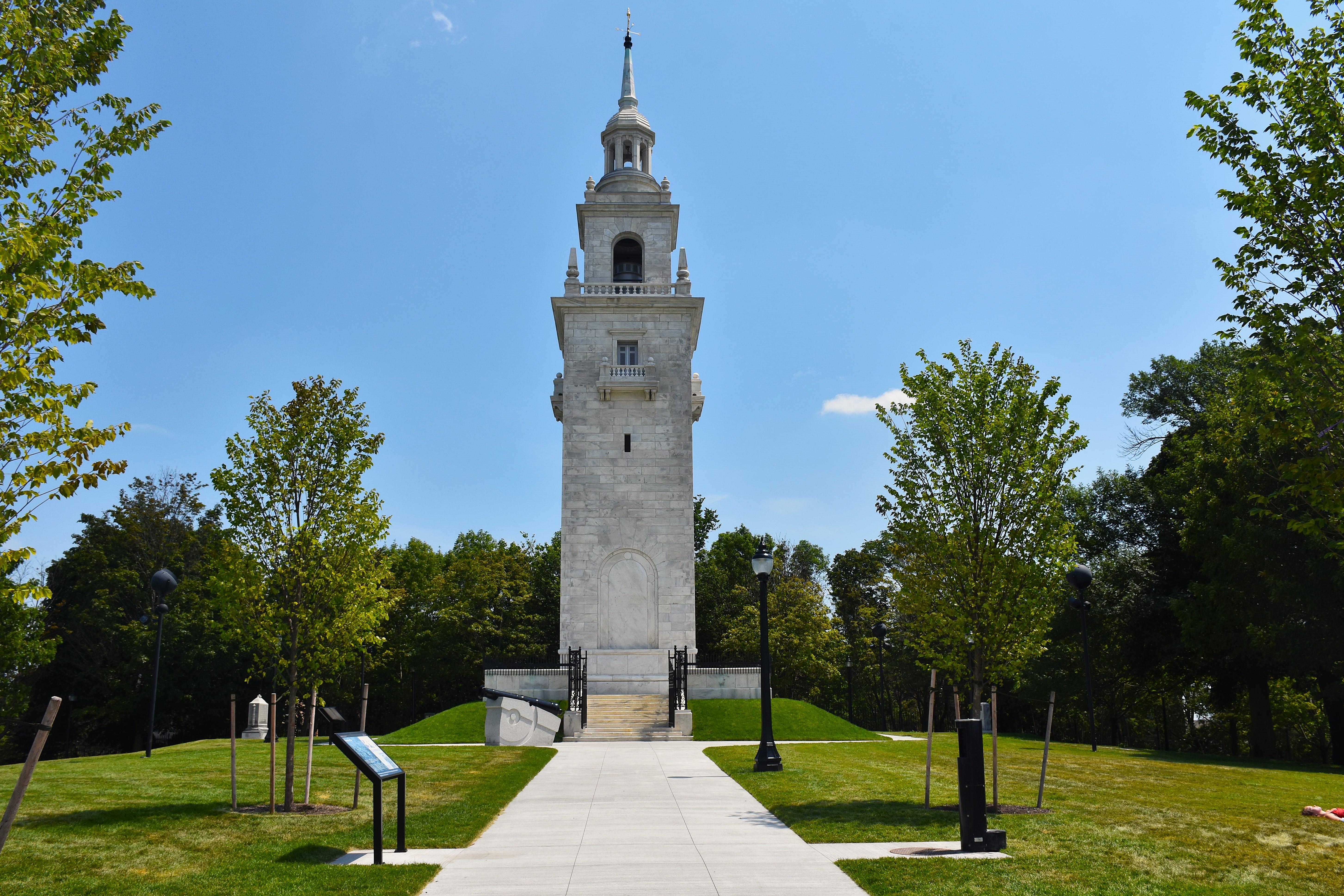 A white marble steeple-looking tower stands amongst trees in a community park.