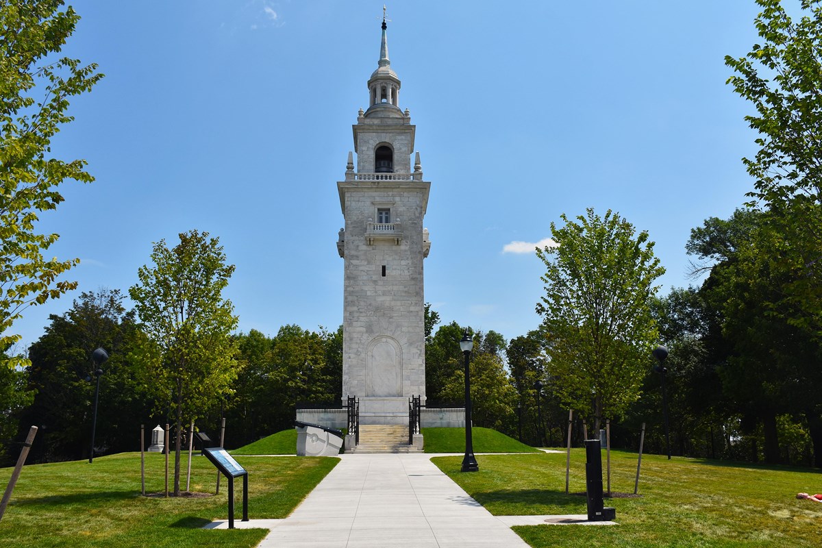 A white marble steeple-looking tower stands amongst trees in a community park.