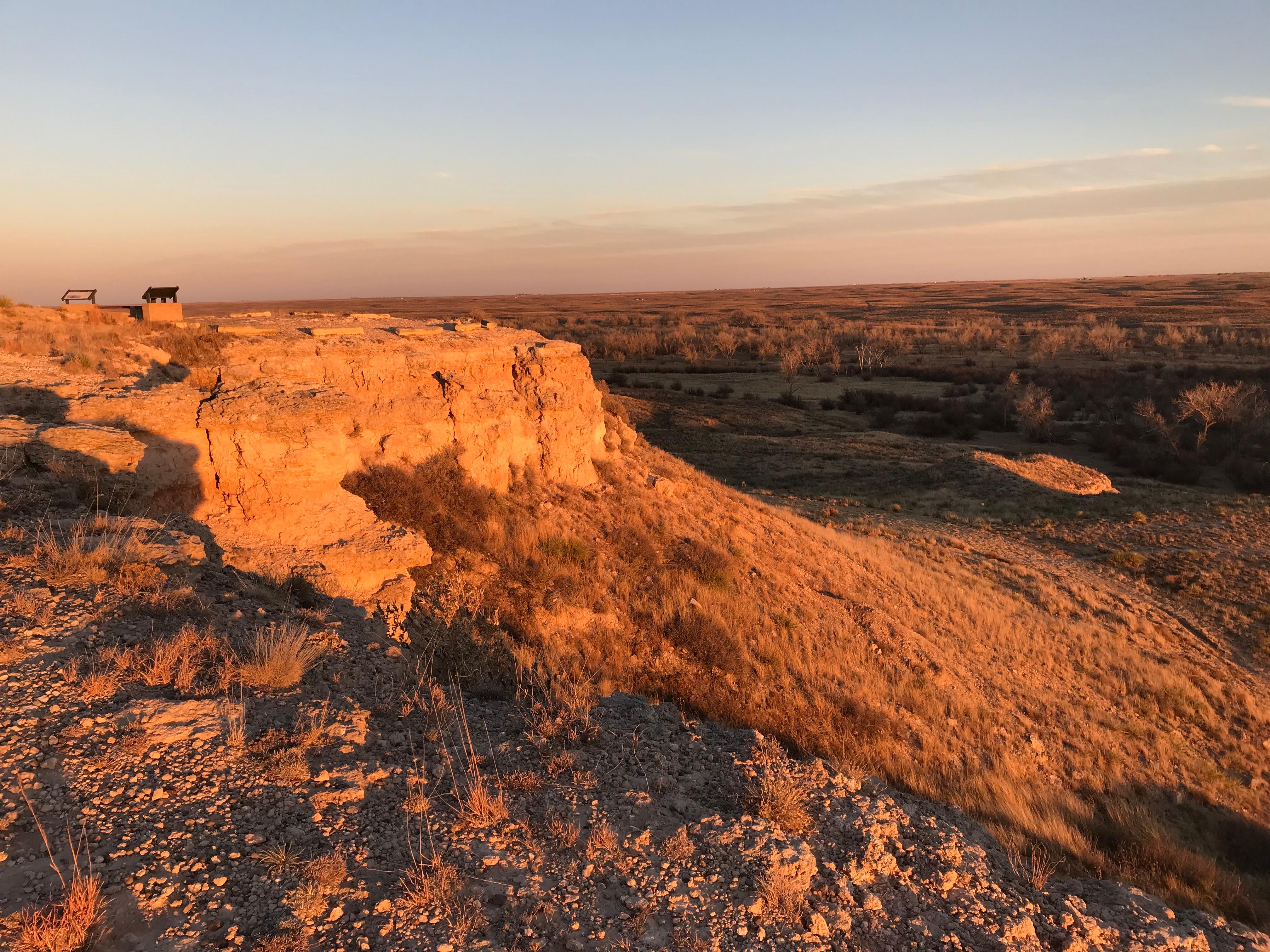 Warm light from the setting sun gives an orange glow to a rock bluff overlooking a grassland.