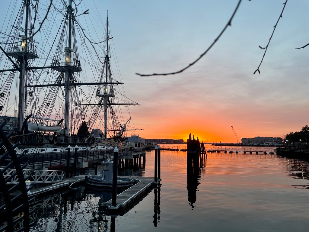 A three-masted ship sits in still water as the sun sets over Boston Harbor.