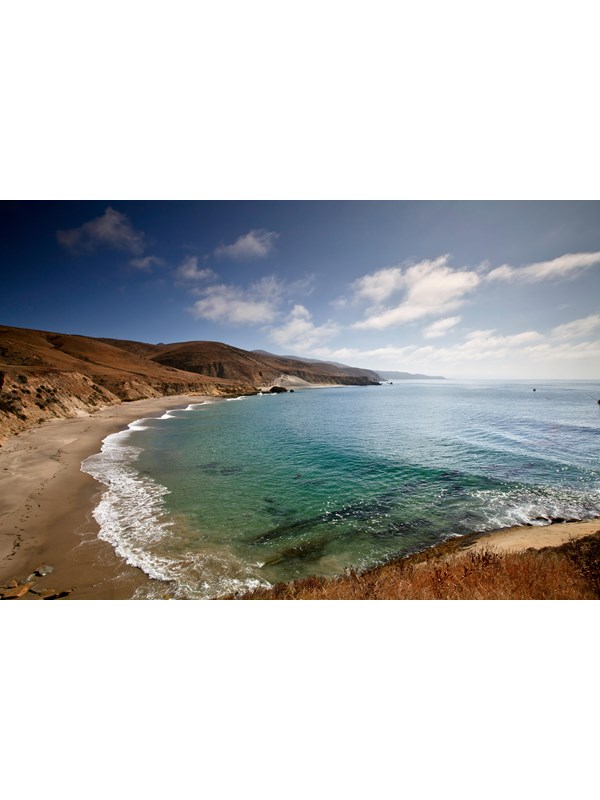 Island coastline with tan sand beach leading out to rolling hills in the distance with green water.