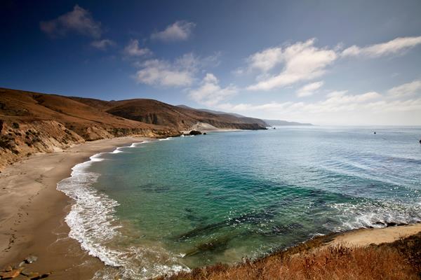 Island coastline with tan sand beach leading out to rolling hills in the distance with green water.