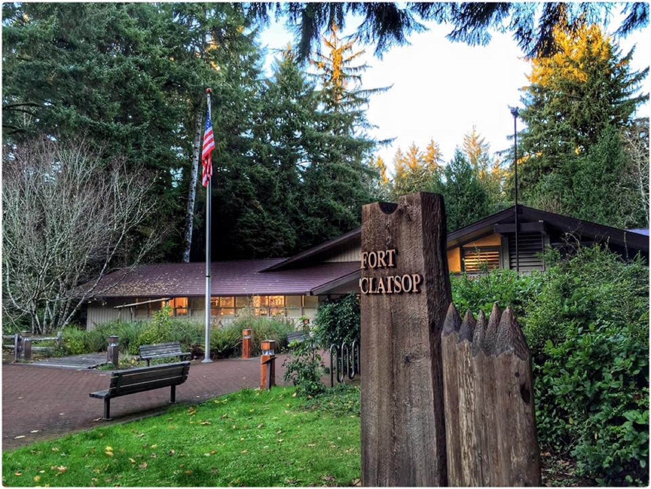A wooden rectangular sign reading Fort Clatsop eclipsing a wide one story building and flag pole