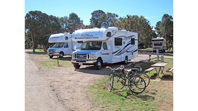 Three RVs in pull-through sites with picnic tables. Trees form the background.