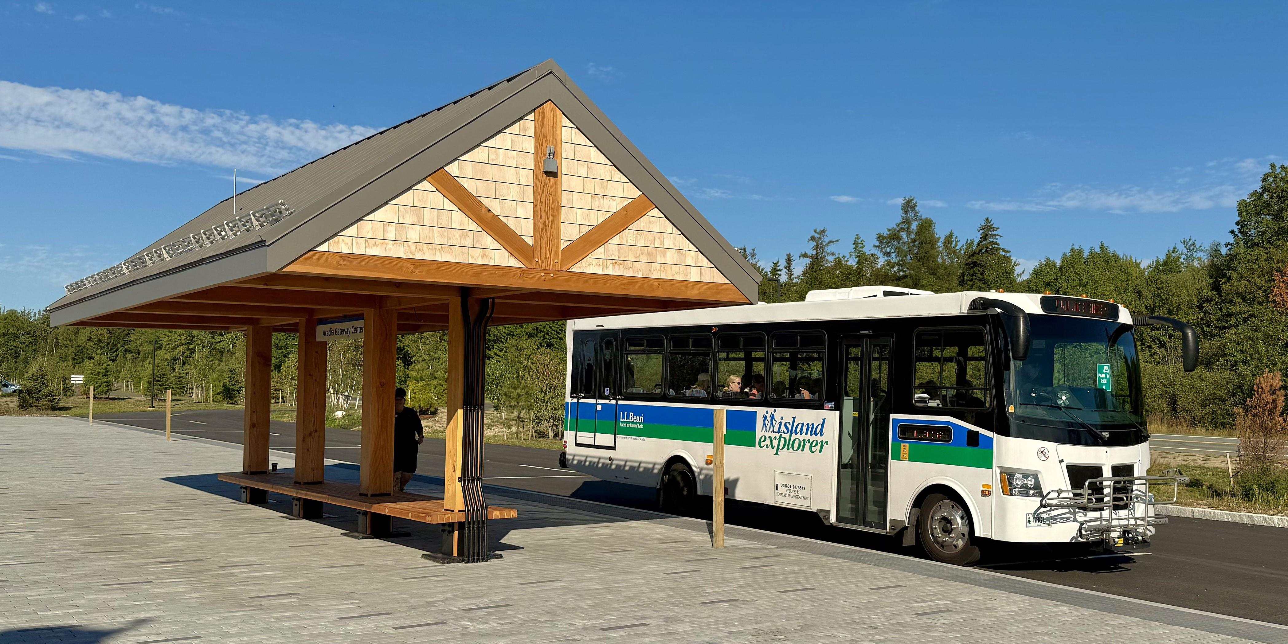 A covered outdoor pavilion with benches near a roadway with a parked an Island Explorer shuttle bus