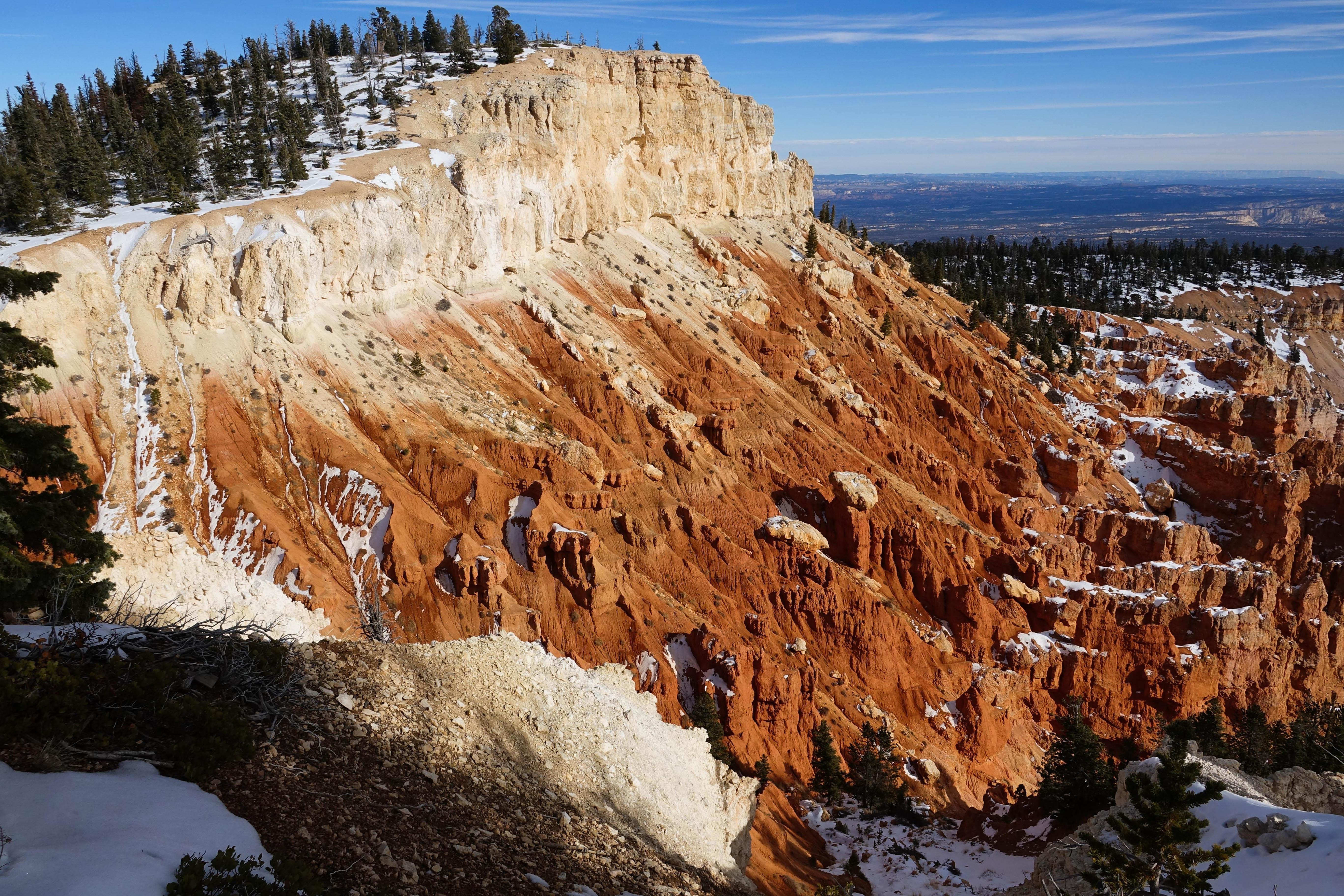 Red and white rock cliffs lightly dusted with snow with a forest atop them and a long view beyond