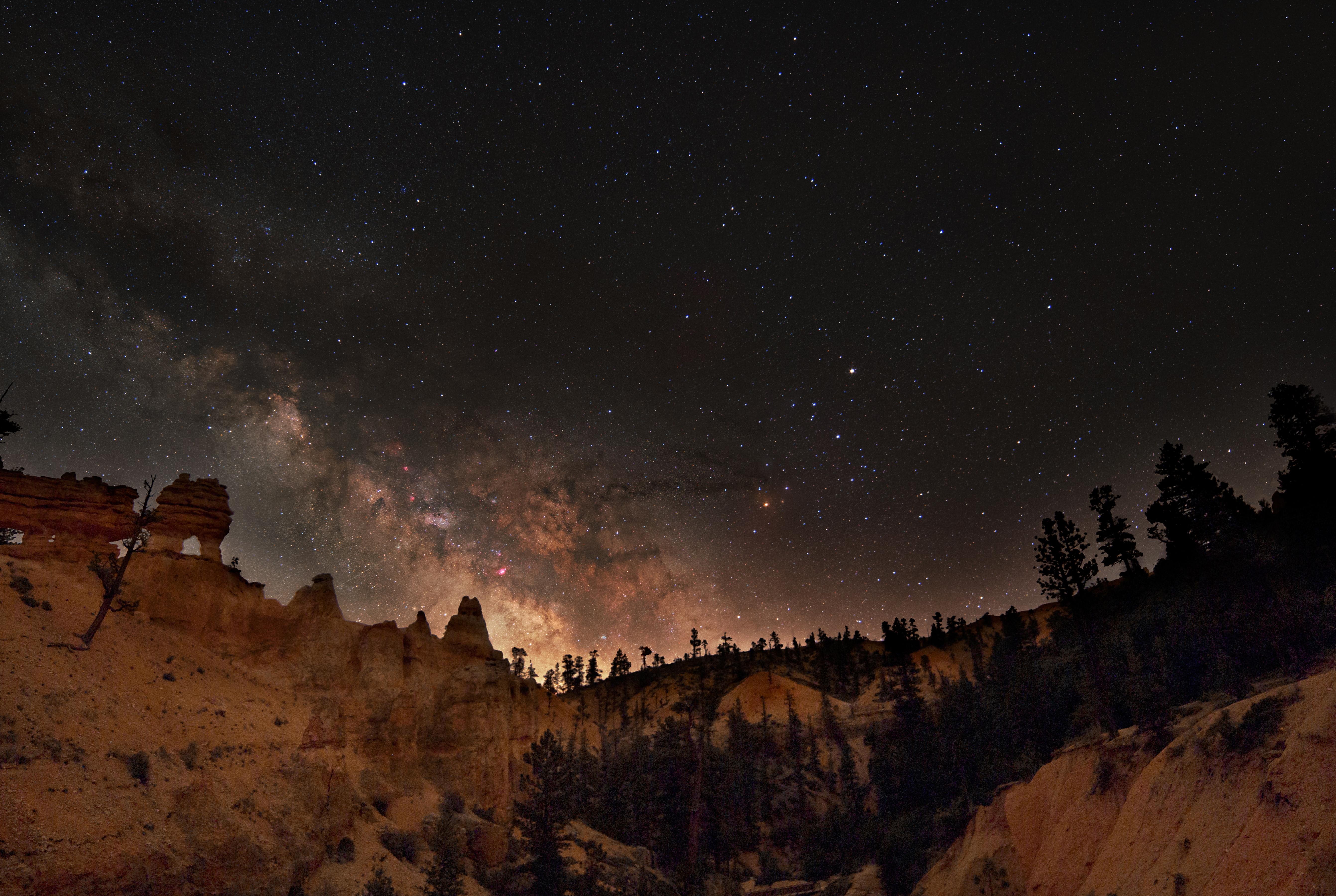 The center of the Milky Way galaxy is seen rising above a horizon of forest and red rock spires