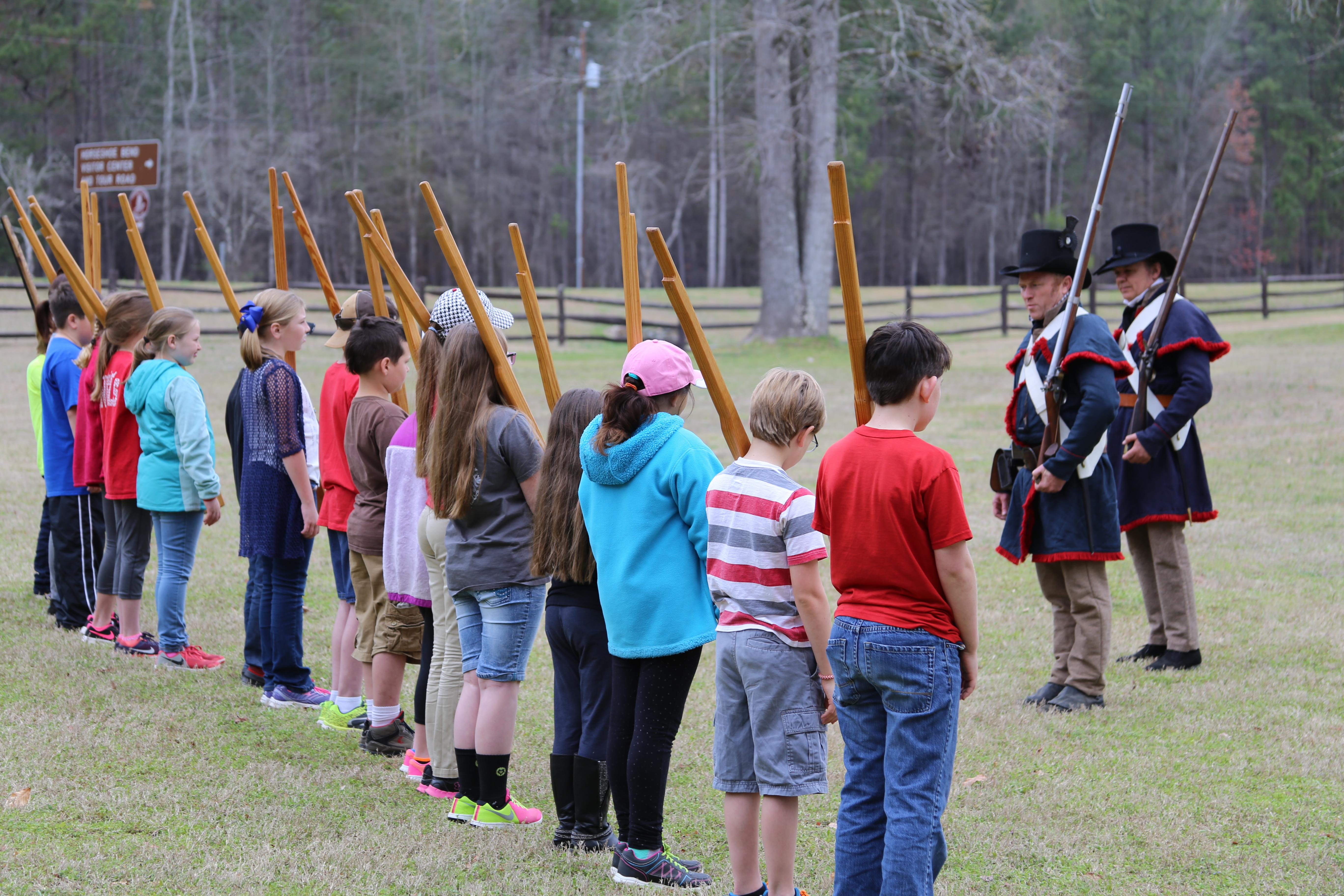 Two rangers dressed in 1812 clothing face a line of children with wooden muskets