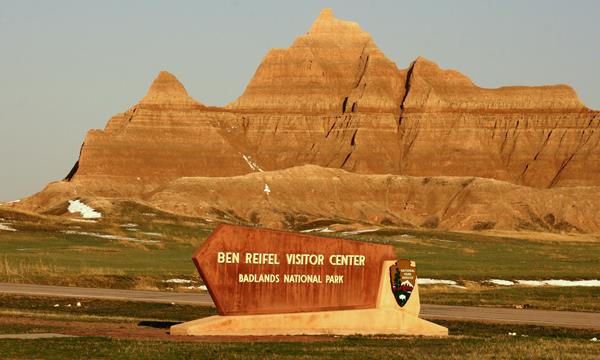 Layered and jagged badlands butte in background with Ben Reifel Visitor Center welcome sign.