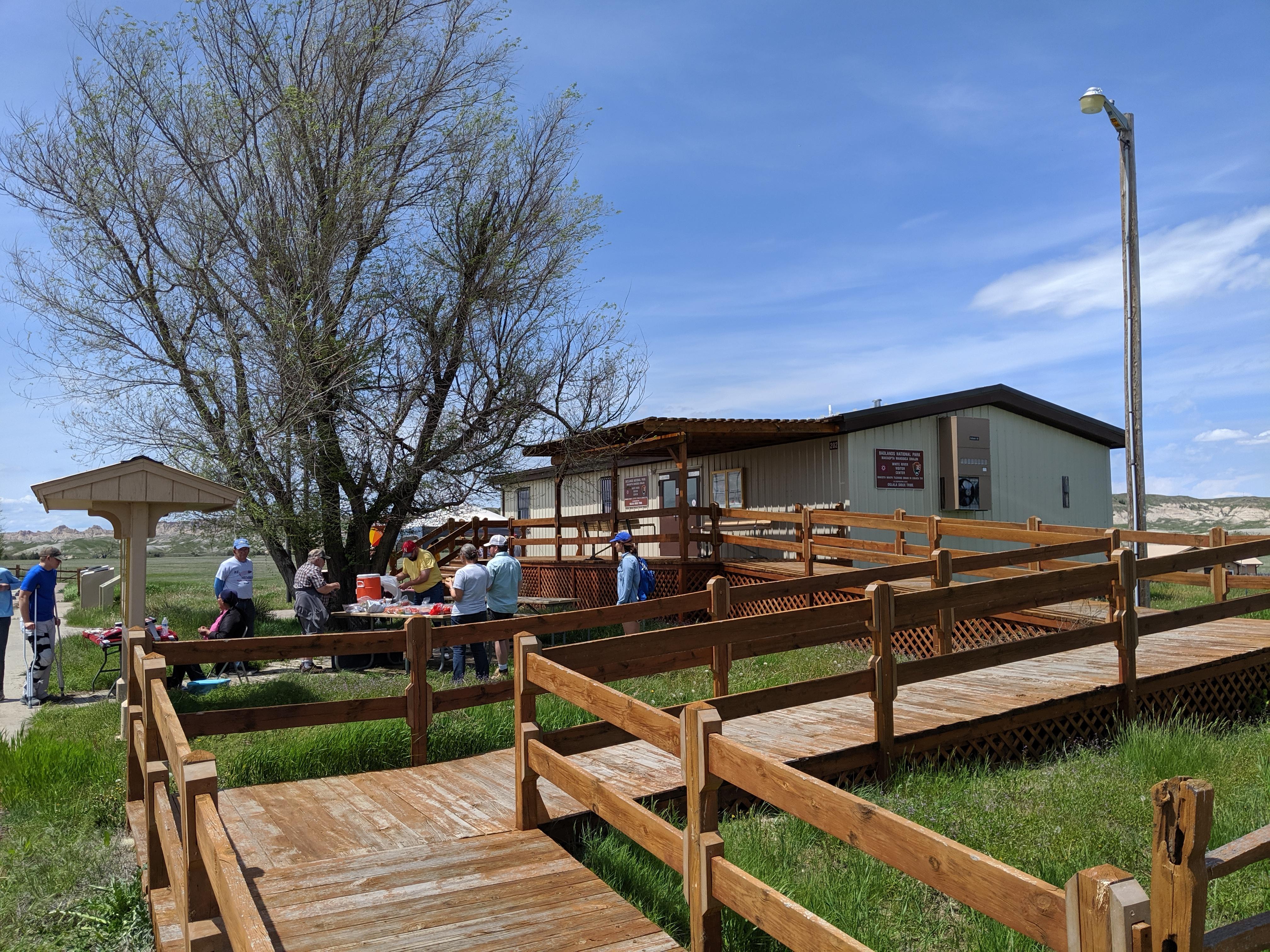 Double-wide trailer with wooden boardwalk ascending to front door under a blue sky.