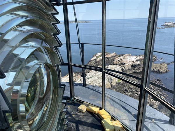 A lighthouse lens to the left, and to the right is a window looking out over a rocky shoreline.