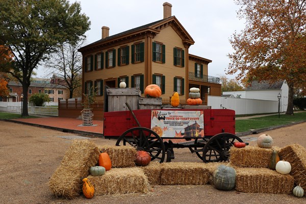 A red cart surrounded by pumpkins and hay bales with the Lincoln Home in the background.