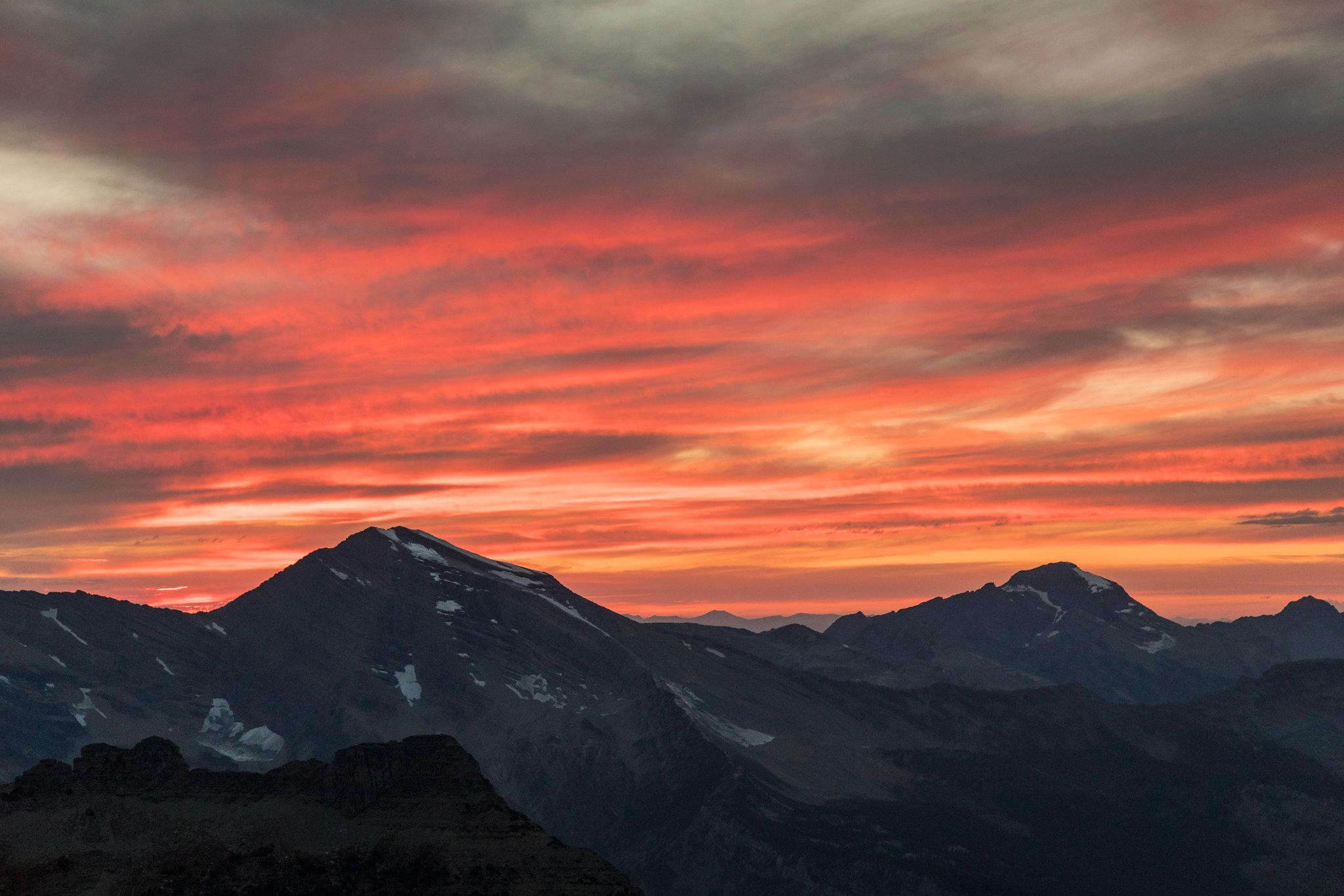 Clouds of orange and red sit above dark-gray mountains; snow dots the mountain peaks.