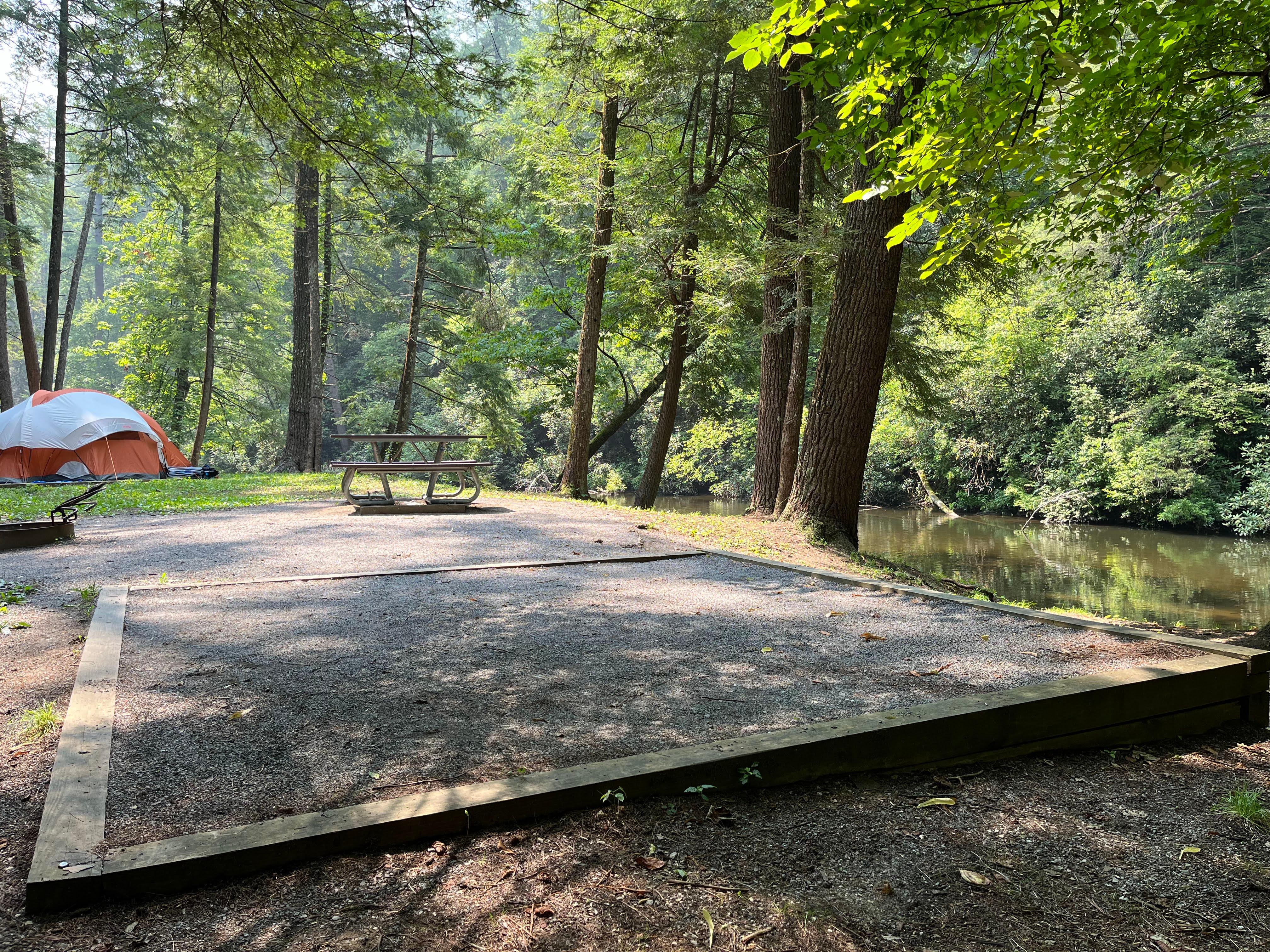 A square tent pad at a waterside site with a picnic table and grill, near an orange and white tent.