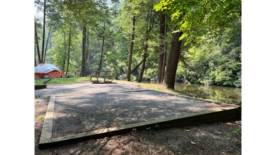 A square tent pad at a waterside site with a picnic table and grill, near an orange and white tent.