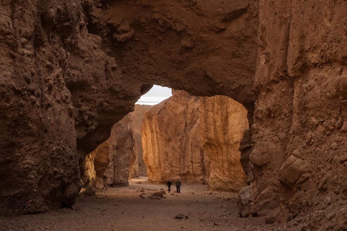 A natural rock arch spans a narrow desert canyon. The viewer stands in deep shade beneath the arch.