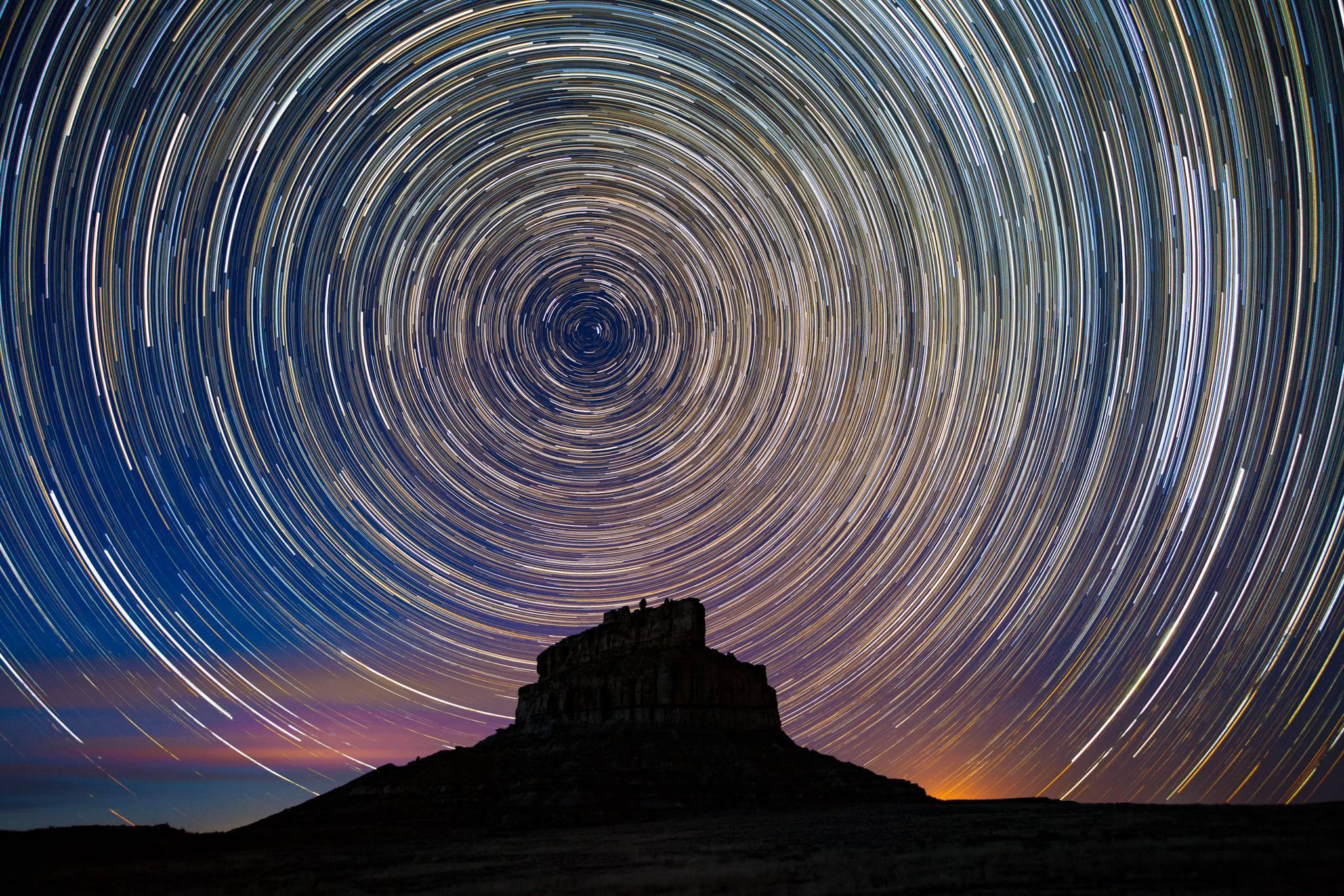Fajada Butte's silhoutte shown under a timelapse image of stars appearing in a vibrant circle above.