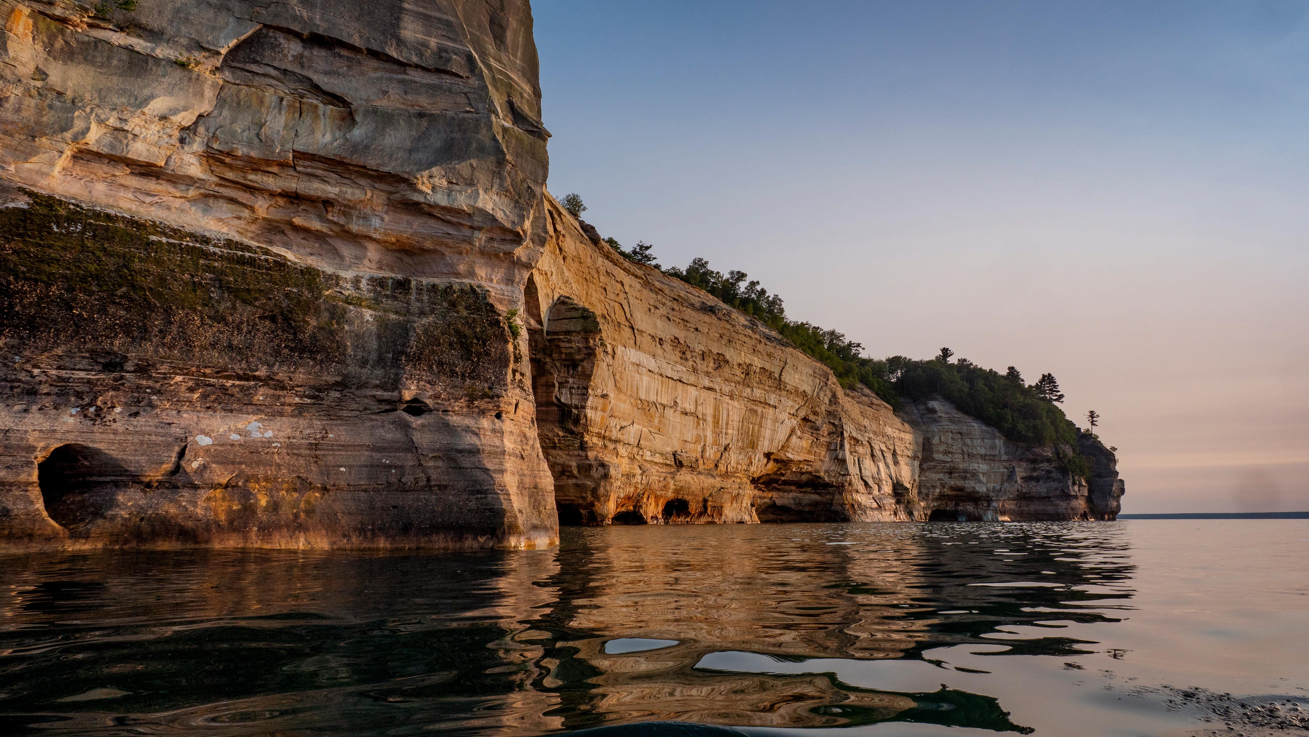 Sandstone cliffs with streaks of brown, red, and orange rise out of Lake Superior at sunset.